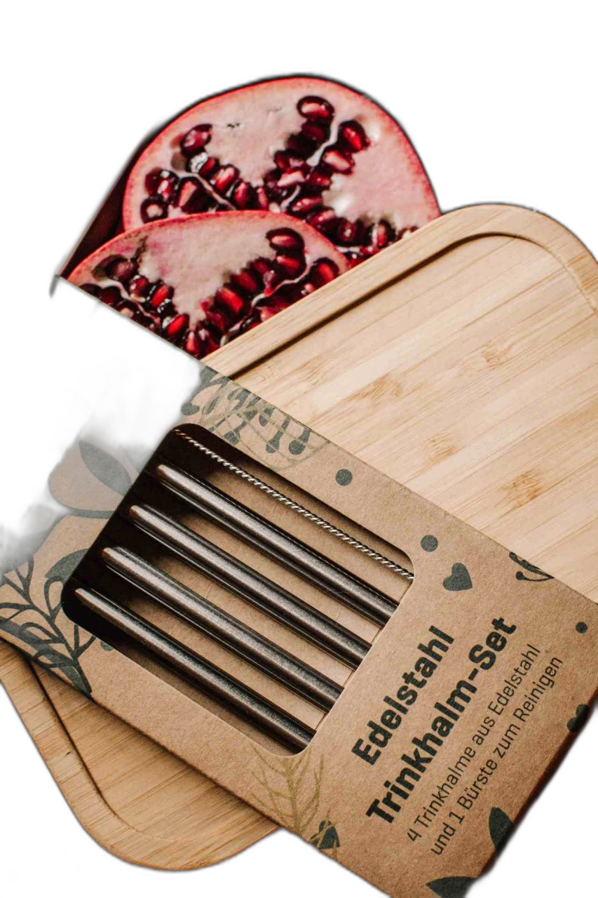 Edelstahl Trinkhalm-Set in Kartonverpackung auf einem Bambusdeckel, im Hintergrund eine geöffnete Metalldose mit frischen Granatapfelhälften und Blaubeeren auf Holztisch.